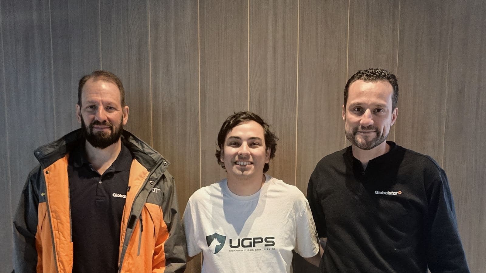 De izquierda a derecha, el director comercial de Globalstar para Latinoamérica, con chaqueta naranja y gris; el CEO de UGPS, Patricio Soto, con polera blanca de UGPS; y Alessandro Allgayer, de Globalstar, con polerón negro, de pie frente a un muro de madera.